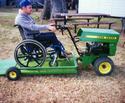 Seen immediately ahead of a stationary access ramp is the wheelchair-accommodating, two-wheeled platform thatâ€™s attached behind the de-tinedâ€™ JD roto-tiller being operated by an adult male.