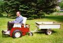 An adult male aboard the chair thatâ€™s pulling a small trailer; the gasoline engine is behind the seat, the large drive wheels are toward the front, and a single swivel wheel is in back.