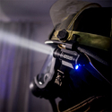 Welding helmet with small flashlight fastened next to left ear and shining forward in dark room.