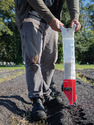 Person from waist down standing in garden row using clear tube with seeds in it and red planting end on tube to jab seeds into row hill.