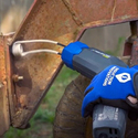 Blue-gloved hand  holding gray & blue pistol-looking heat induction tool that has 2 heavy wires coming out of the pistol muzzle with a coil on the far end holding against a seized nut & bolt